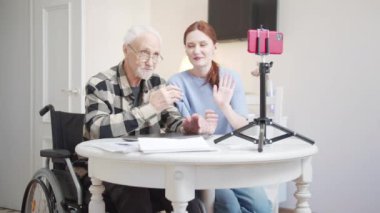 Tekerlekli sandalyede bir adam ve genç bir kadın oturma odasındaki masanın önündeler. Masanın üzerinde tripodun üzerinde bir akıllı telefon var. Seyircilerin sorularını yanıtlarlar.