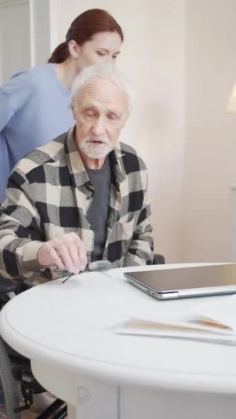 若い看護師が車椅子で老人の世話をする 彼女は彼をテーブルにピンチし 彼がコンピュータをオンにするのを助けます 高品質のフルHd映像 — ストック動画