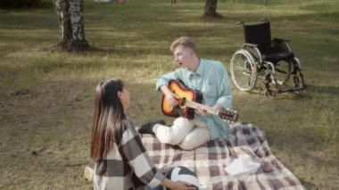 Elinde gitar olan engelli genç bir adam ekose üzerinde oturuyor ve kız arkadaşı başını sallayıp biraz dans ederken şarkılar söylüyor. Yüksek kaliteli FullHD görüntüler