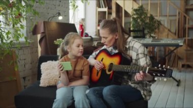 Aile hobileri. Bir kadın gitar dersi alıyor. Küçük çocuğu yardım ediyor ve telefondaki akorları gösteriyor. Yüksek kalite 4k görüntü