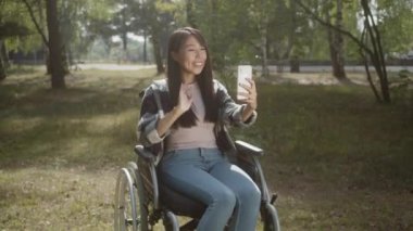 Ekose gömlekli engelli bir Asyalı kız tekerlekli sandalyesinde oturuyor ve akıllı telefonunun ekranına bakıyor, el sallıyor ve arkadaşlarıyla konuşuyor. Yüksek kalite 4k görüntü