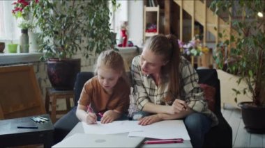 Çocuk annesiyle ev ödevi yapıyor. Bir kadın kanepeye oturur ve kızın defterine ne yazdığını izler.