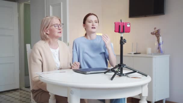 소녀와 할머니가 삼각대 전화기 탁자에 있습니다 여자는 버튼을 누르고 비디오를 — 비디오