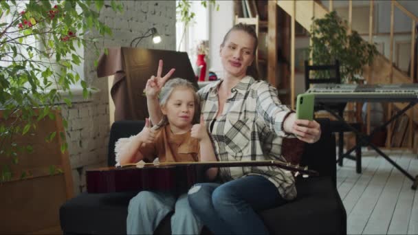 一个女人用智能手机给自己和女儿拍了一张照片 他们彼此吹喇叭 并展示吉他 他们看着手机屏幕 给照片打分 他们玩得很开心 — 图库视频影像