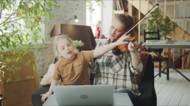Çocuk keman çalmada anneye yardım ediyor. Dizüstü bilgisayarda ders izleyen ve bir kadını düzelten bir kız. Yüksek kalite 4k görüntü