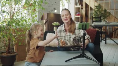 Bir kadın ve çocuğu sıcak bir oturma odasında oturuyorlar. Önlerindeki masada satranç taşları ve tripodun üzerinde bir telefon var. Bir kız, annesiyle birlikte cep telefonunun düğmesine basıyor.