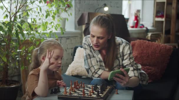 女性と彼女の娘はチェスのゲームをプレイし ビデオチュートリアルで携帯電話を使用します — ストック動画