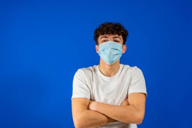 Steril yüz maskeli genç adam mavi arka plan stüdyo resminde izole edilmiş bir şekilde poz veriyor. Salgın hızla yayılan koronavirüs 2019 ncov sarar covid-19 grip virüsü konsepti. El ele tutuşarak..