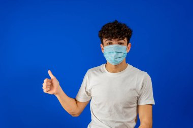 Gibi. Cerrahi maske takmış tatmin olmuş genç bir adamın portresi başparmağıyla ayakta kameraya bakıp gülümsüyor. Mavi arka planda kapalı stüdyo çekimleri.