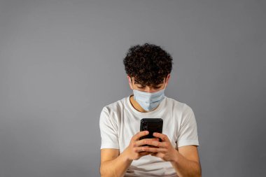 Steril yüz maskeli genç adam gri arka plan stüdyo resmine izole edilmiş poz veriyor. Salgın Coronavirüs 2019-ncov sarars covid-19 grip virüsü konsepti. Cep telefonu kullanmak, SMS iletisi yazmak