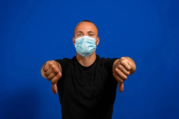 Νεαρός καυκάσιος άνδρας με κοντά μαλλιά που φοράει σπασμένη ιατρική μάσκα πάνω από απομονωμένο μπλε φόντο που αναστατώνεται δείχνοντας τον αντίχειρα κάτω με δύο χέρια. Αντιπαθητική έννοια. — Φωτογραφία Αρχείου