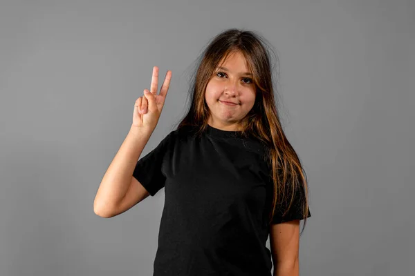 Gesture and happy people concept - uśmiechnięta dziewczynka w czarnym t-shircie pokazująca gest pokoju z palcami na szarym tle studia — Zdjęcie stockowe