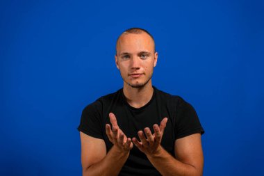 Mavi izole edilmiş arka planda sıradan siyah gömlek giyen genç, yakışıklı İspanyol adam avuçlarını birbirine değdirirken ya da el hareketi yaparken gülümsüyor. Tutun ve koruyun.