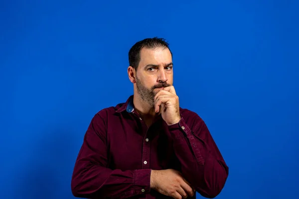 Retrato de jovem homem pensativo com a mão perto do rosto com expressão pensativa contra fundo azul no estúdio — Fotografia de Stock