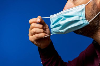 Mavi arka planda izole edilmiş cerrahi maskesini çıkartan İspanyol adamın yüzü Coronavirus 'un neden olduğu yeni normal durumun yarattığı maskeden kurtulur.