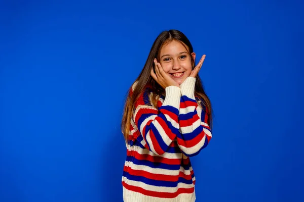 Linda menina pré-adolescente espanhola vestida com uma camisola listrada com as palmas das mãos em seu rosto em um gesto adorável, isolado no fundo do estúdio azul Imagem De Stock