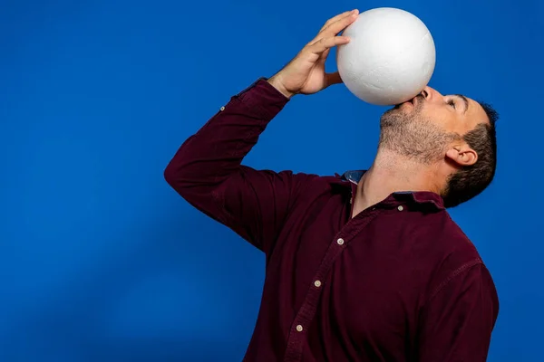 Primer plano del hombre caucásico emocional jugando al fútbol golpeando la pelota con la boca sobre un fondo azul aislado. Fútbol, deporte, expresión facial, emociones humanas, concepto de estilo de vida saludable. Espacio de copia — Foto de Stock