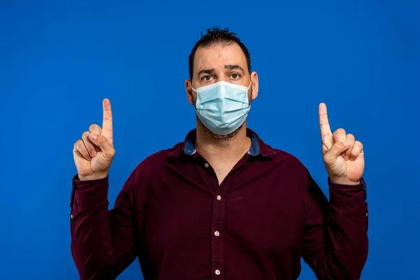 Νεαρός με αποστειρωμένη μάσκα που ποζάρει απομονωμένος σε μπλε φόντο. Η επιδημιολογική πανδημία εξαπλώνεται γρήγορα coronavirus 2019-ncov sars covid-19 έννοια του ιού της γρίπης. Δείκτες σημείου επάνω — Φωτογραφία Αρχείου