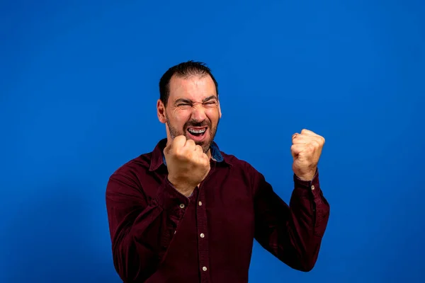 Homem hispânico com barba celebrando energeticamente com punhos apertados, isolado no fundo do estúdio azul. — Fotografia de Stock