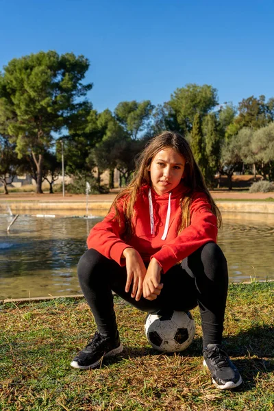 Beautiful preteen girl sitting on top of a soccer ball in a beautiful park with natural grass. She is resting after exercising with the ball. — Stock Photo, Image