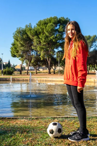 Preteen Kaukasische atleet meisje poseren met haar voetbal in een prachtig park met een fontein met water op de achtergrond — Stockfoto