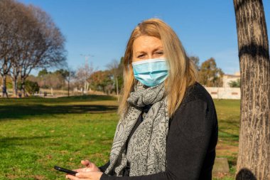 A woman using a smartphone wears a medical face mask to avoid the spread coronavirus on a park. A close-up photo of a woman with a surgical mask on the face against COVID-19.