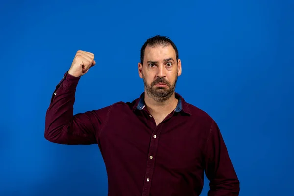 Spanish bearded man in a purple shirt pulling out muscle with a puzzled face as he has lost his muscle power from when he was young. Disappointment concept. — Fotografia de Stock