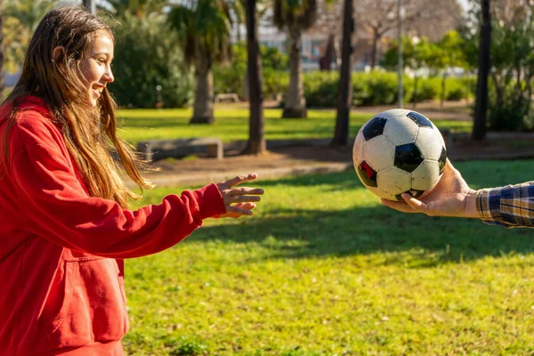 Mans mano que ofrece una pelota de fútbol a una niña preadolescente bastante sonriente para jugar fútbol en el parque. La chica quiere practicar para ser un mejor jugador de fútbol. — Foto de Stock