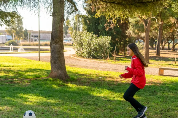 Krásná holčička hraje fotbal v pěkném parku s přirozenou trávou za slunečného zimního dne. Cvičení a zdravý život koncept. — Stock fotografie