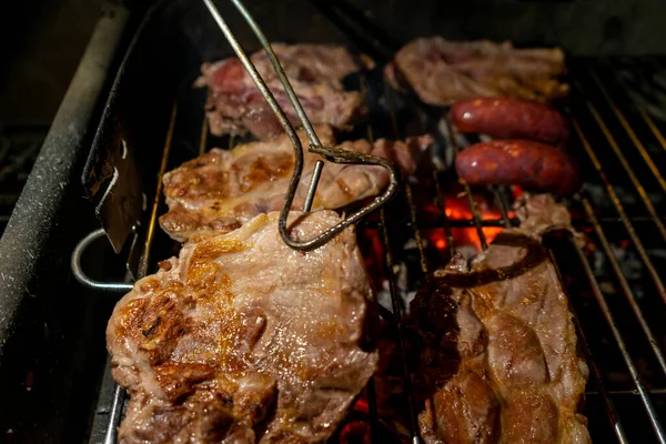 Carne de cerdo a la parrilla en la barbacoa con pinzas de metal utensilios en la parrilla en llamas barbacoa con humo. Comida al aire libre. Enfoque selectivo. —  Fotos de Stock