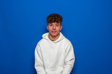 Kıvırcık saçlı, beyaz kazaklı, mavi bir arka plandan kameraya bakan ciddi yakışıklı, beyaz bir beyaz adam..