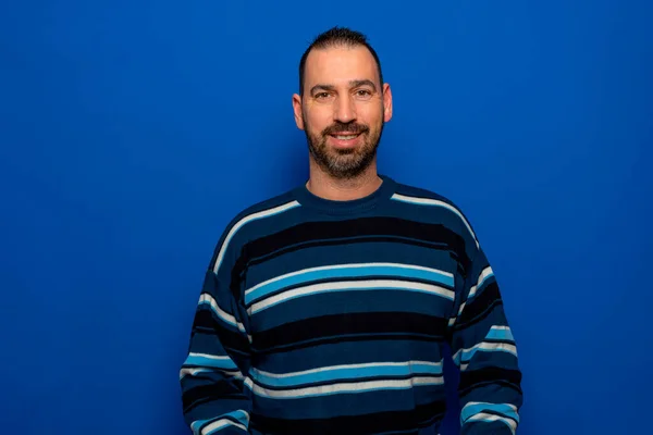 Retrato de um homem espanhol sorridente em seus 40 anos vestindo um pulôver listrado isolado no fundo do estúdio azul. — Fotografia de Stock