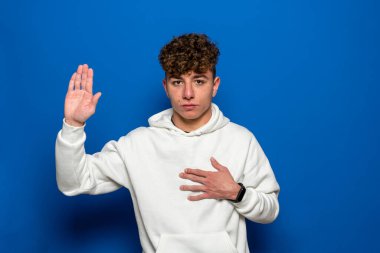Kıvırcık saçlı, mavi arka planda sıradan bir süveter giyen, elleri göğsünde ve avuçları açık yemin eden, sadakat yemini eden genç yakışıklı bir adam..