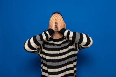 Siyah-beyaz çizgili süveter giyen beyaz bir adam mavi arka planda izole edilmiş bir umutsuzluk jesti ile yüzünü kapatıyordu..