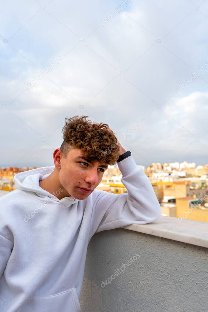 Portrait of smiling young Caucasian man with curly hair dressed in a white sweatshirt looking at the camera with the city skyline in the background