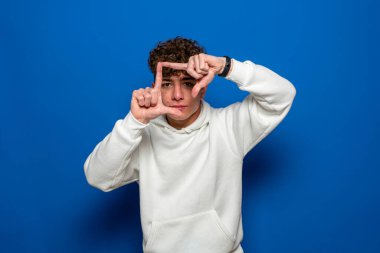 Kıvırcık saçlı, mavi arka planda beyaz kazak giyen, gülümseyen, elleri ve parmakları mutlu yüzlü genç, yakışıklı esmer adam. Yaratıcılık ve fotoğrafçılık kavramı.