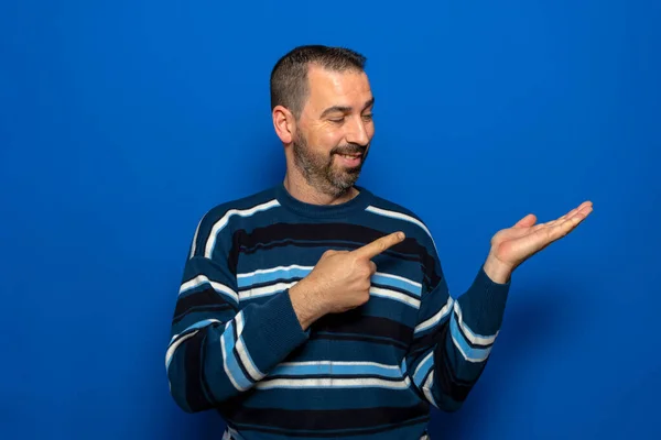 Homem espanhol com barba sobre fundo azul isolado espantado e sorrindo ao apresentar com a mão e apontando o dedo. — Fotografia de Stock