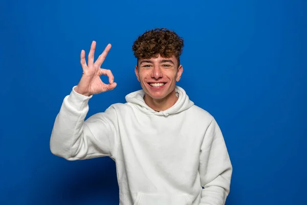 Young caucasian man with curly hair wearing a white sweatshirt standing on white background with a happy face smiling doing an ok sign with his hand isolated on blue studio background. — Stock Photo, Image