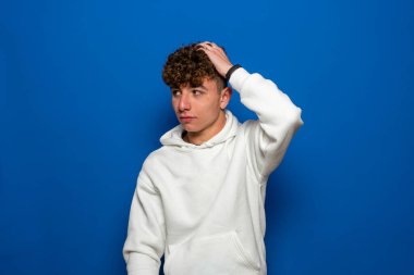 Genç yakışıklı, uzun boylu, zayıf, kahverengi saçlı, mavi arka planda şüpheli bir hareketle başını kaşıyan beyaz adam..