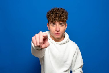 Siyah saçlı, mavi arka planda beyaz kazak giyen genç ve yakışıklı bir adam kameraya kızgın ve sinirli olduğunu gösteriyor..