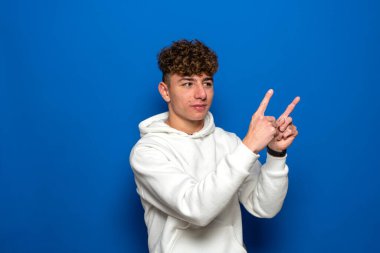 Beyaz kazak giyen, beyaz tişörtlü, mutlu beyaz bir beyaz adamın portresi. Parmak uçlarını kenarı gösteriyor. Fotokopi alanını mavi arka planda bırakıyor.