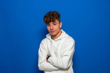 Beyaz kazak giymiş beyaz saçlı, beyaz saçlı, kollarını mavi bir stüdyonun arka planında çapraz pozlar veren beyaz bir genç..