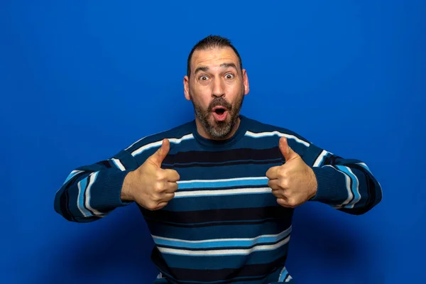 Homem bonito jovem vestindo camisola casual em pé sobre isolado sinal de sucesso fundo azul fazendo gesto positivo com a mão, polegares para cima sorrindo e feliz. Expressão surpreendente. — Fotografia de Stock