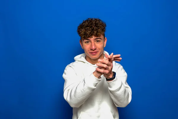 Jeune homme brune beau aux cheveux bouclés portant un sweat-shirt blanc applaudissant les mains sur fond bleu et applaudissant heureux et joyeux, souriant fiers mains ensemble. — Photo