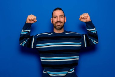 Beyaz, mutlu, tıraşsız, güçlü spor spor spor, 40 'lı yaşlarda çizgili süveter giyen, arka planda mavi renkli stüdyo portresi üzerinde duran pazı kaslarını gösteren..