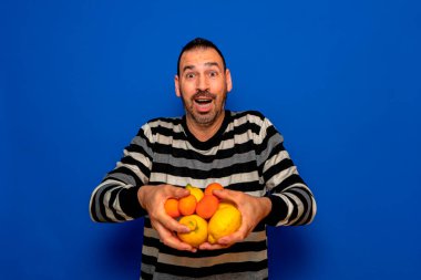 Çizgili süveter giyen İspanyol sakallı adam mavi stüdyo arka planında elinde tuttuğu portakal ve limonları heyecanla sunuyor..