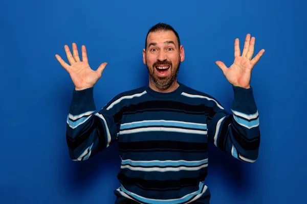 Portret van een opgewonden casual man die met opgeheven handen naar camera op een blauwe achtergrond kijkt. — Stockfoto