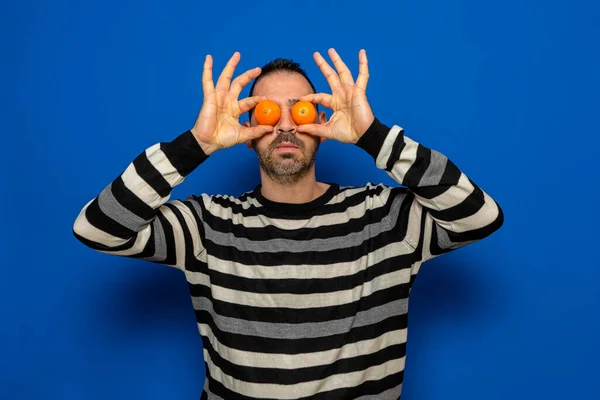 Homem barbudo hispânico segurando duas laranjas perto de seus olhos imitando óculos isolados no fundo do estúdio azul. Conceito divertido — Fotografia de Stock