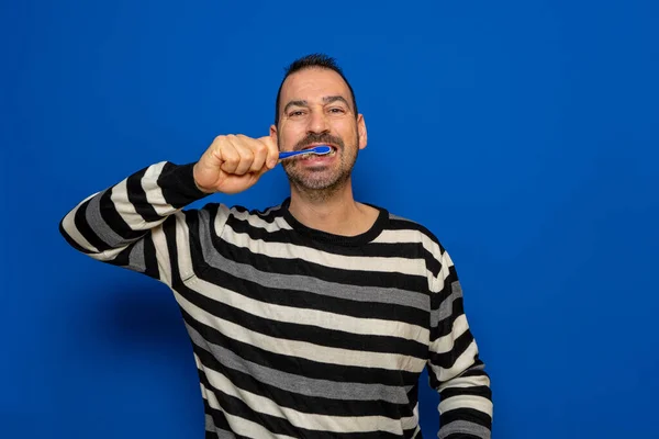 Homem de barba hispânica escovando os dentes isolados no fundo azul, ele tem dentes amarelos e gengivas inchadas porque negligenciou sua higiene oral. — Fotografia de Stock