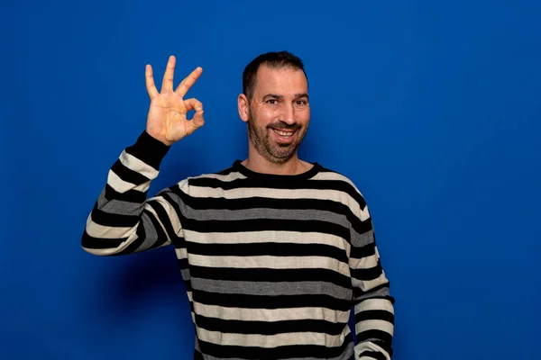 Homem barbudo espanhol em seus 40 anos vestindo um pulôver listrado fazendo o gesto ok enquanto sorri, isolado sobre um fundo de estúdio azul — Fotografia de Stock